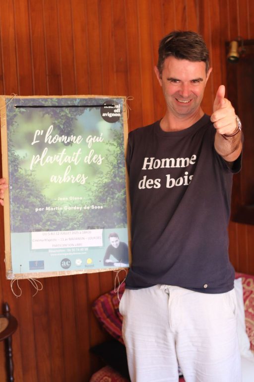 un homme avec un Tshirt où est écrit Homme des bois le pouce levé et un grand sourire. Il tient l'affiche du spectacle