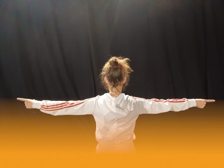 une femme avec veste de survêtement blanc les bras à l'horizontal de dos avec toile noire en fond d'image