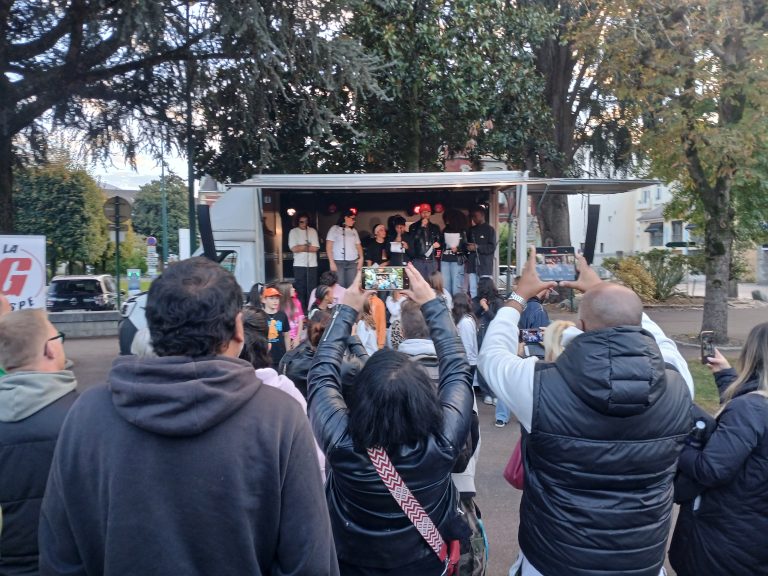 Un camion aménagé en salle de concert avec des jeunes chantant dedans et des personnes de dos prenant des photos du concert avec leur smartphone