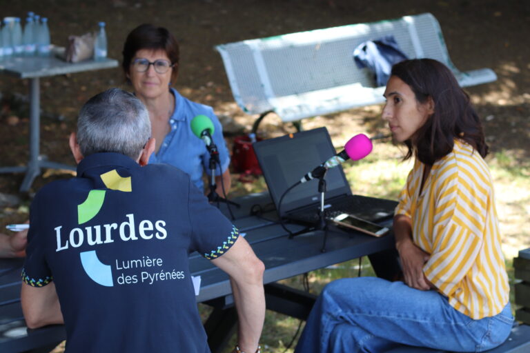 Plateau radio sous les arbres avec un homme de dos portant un tshirt "Lourdes, ville lumière" et deux femmes au micro