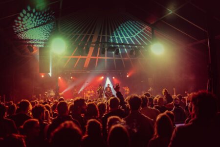 La salle de spectacle sur une teinte rougoyante avec la foule de dos au premier plan et la scène des artistes au fond.